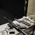 Assorted firearms arranged on a wooden table in an indoor shooting range setting.